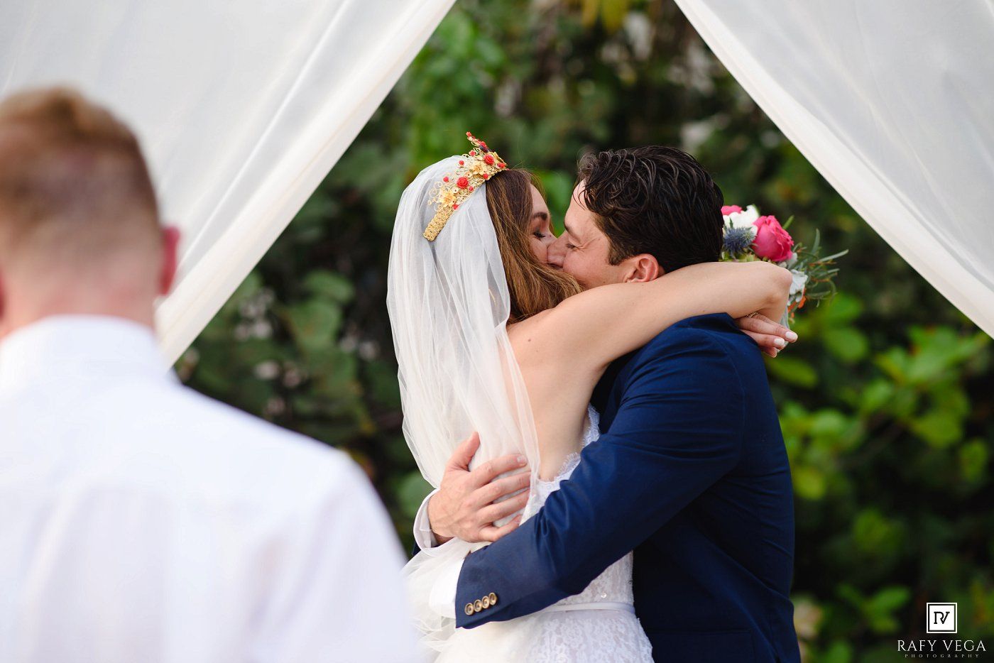 Bride and groom share their first kiss under a white draped arch at an outdoor wedding ceremony.