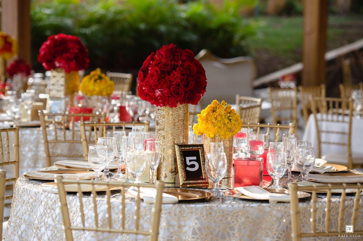 Elegant wedding reception tables with red and yellow floral centerpieces, gold chiavari chairs, and white lace tablecloths.