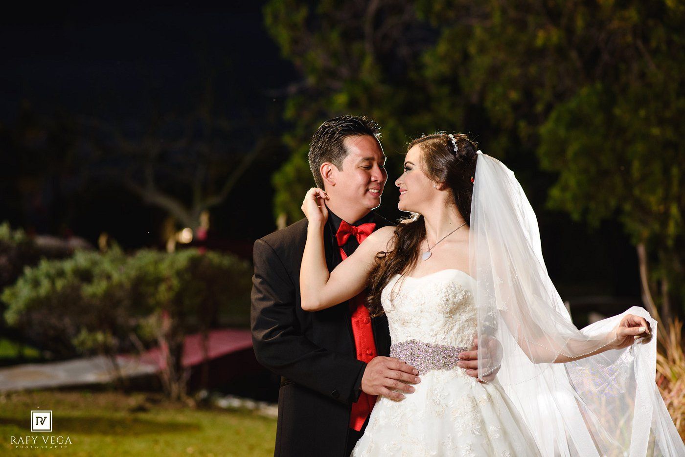 Bride in white lace gown and veil gazes at groom in black suit with red tie during romantic outdoor night wedding.