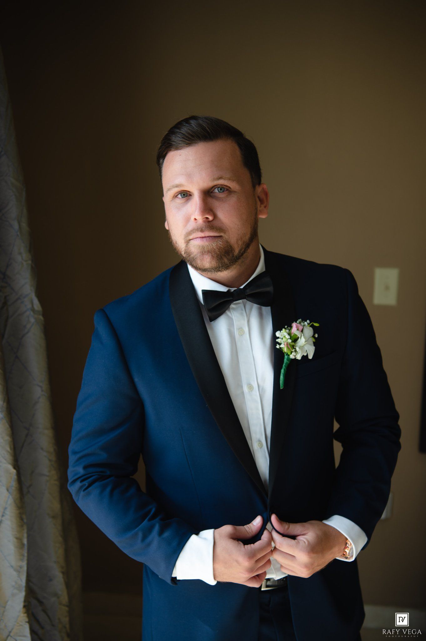 Elegant groom in navy blue tuxedo with black lapels and floral boutonniere posing confidently indoors.