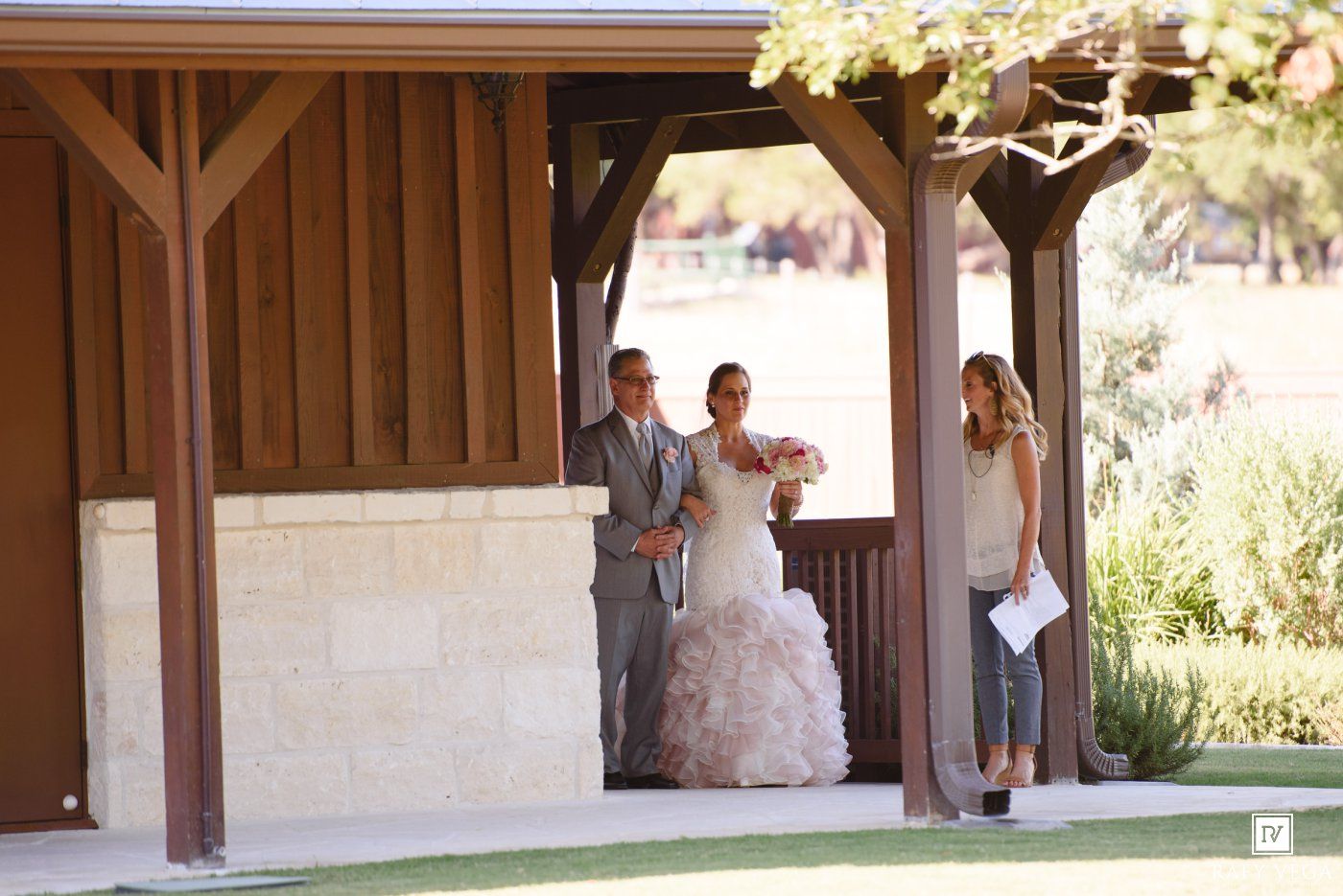 Bride in ruffled gown walks with escorts under wooden pergola at outdoor wedding venue.