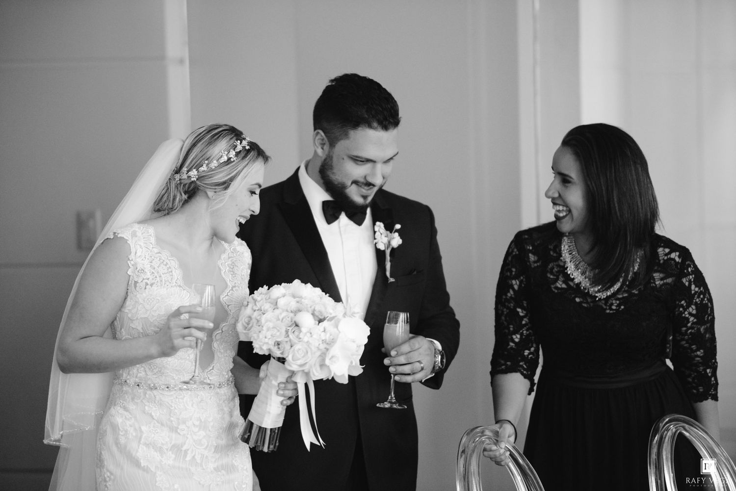 Black and white photo of a smiling bride and groom in wedding attire sharing a toast with a female guest.