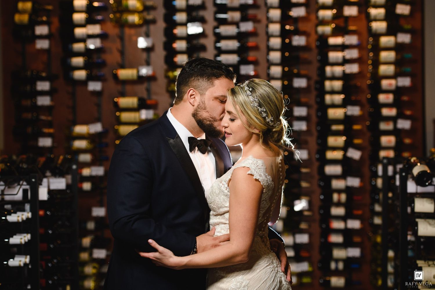 Newlywed couple sharing a romantic kiss in front of an elegant wine cellar wall during their wedding reception.