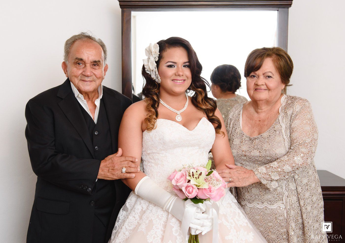 Bride in white lace gown holding pink bouquet poses with elderly man and woman in formal attire before wedding ceremony.