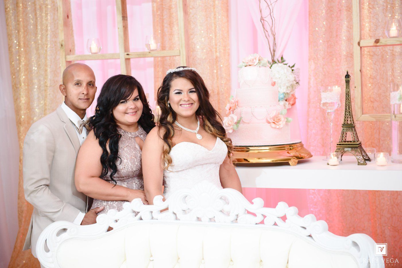 Three people pose together at an elegant quinceañera celebration with a tiered cake and Eiffel Tower decor.