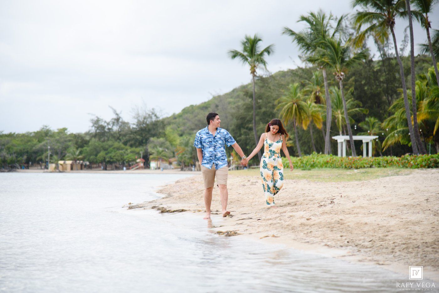 Couple holding hands walking on tropical beach with palm trees and calm water in background.