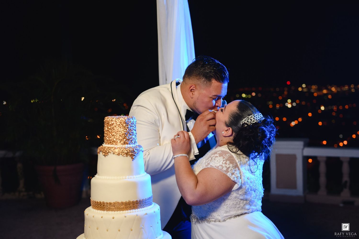 Newlyweds share a kiss beside their elegant four-tier wedding cake at night.