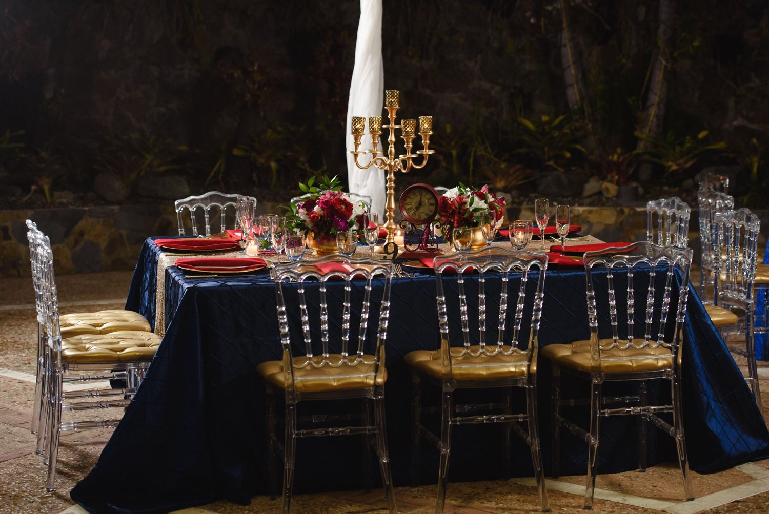 Elegant outdoor dining table with navy blue velvet tablecloth, red napkins, gold candelabra, and acrylic chiavari chairs at night.