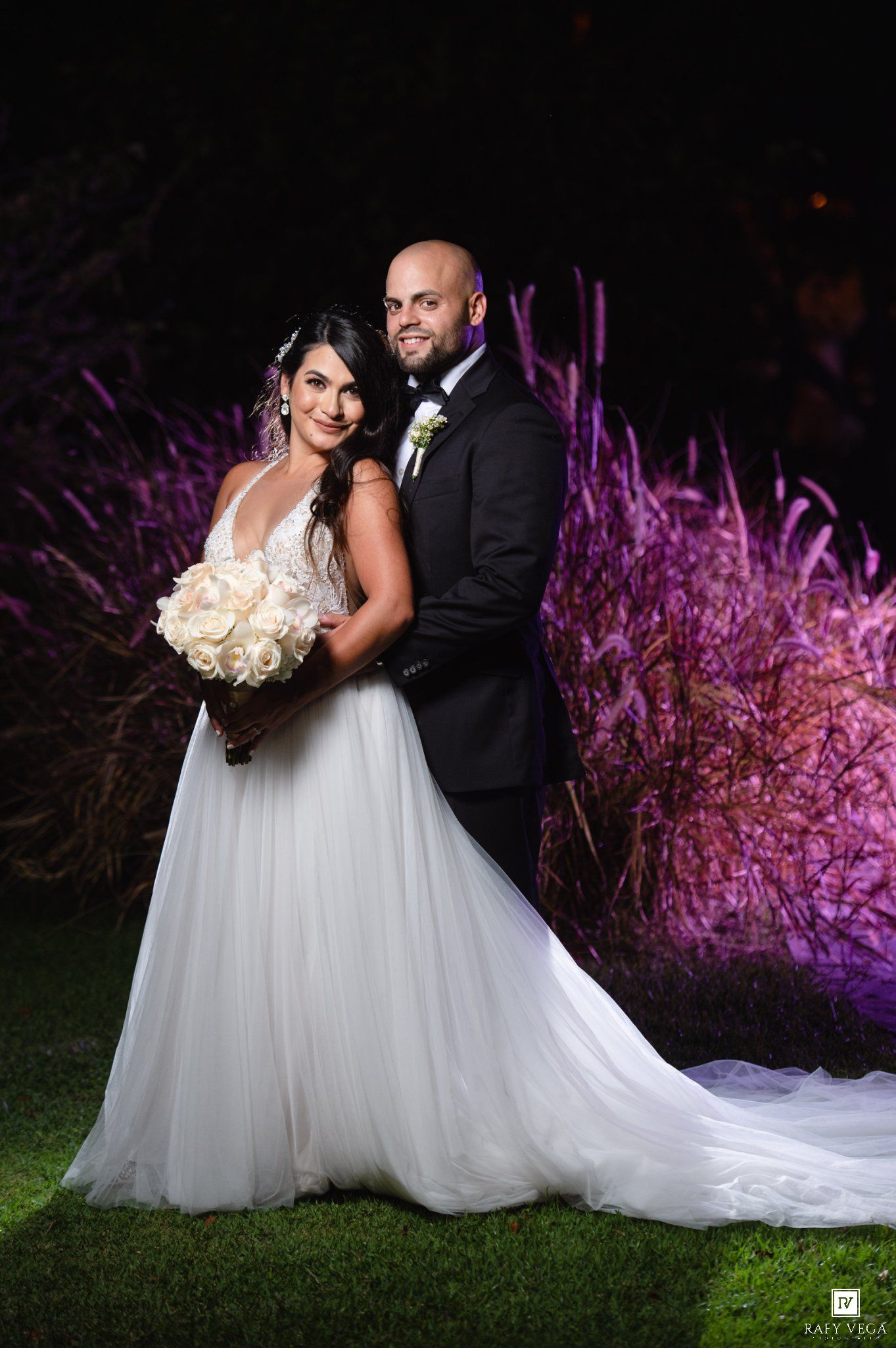 Bride in white gown holding bouquet poses with groom in tuxedo at night wedding with purple lit foliage.
