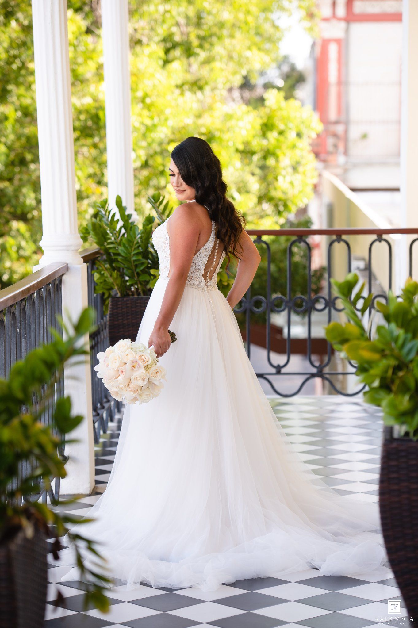Bride in elegant white backless wedding gown holding bouquet on ornate balcony surrounded by lush greenery.