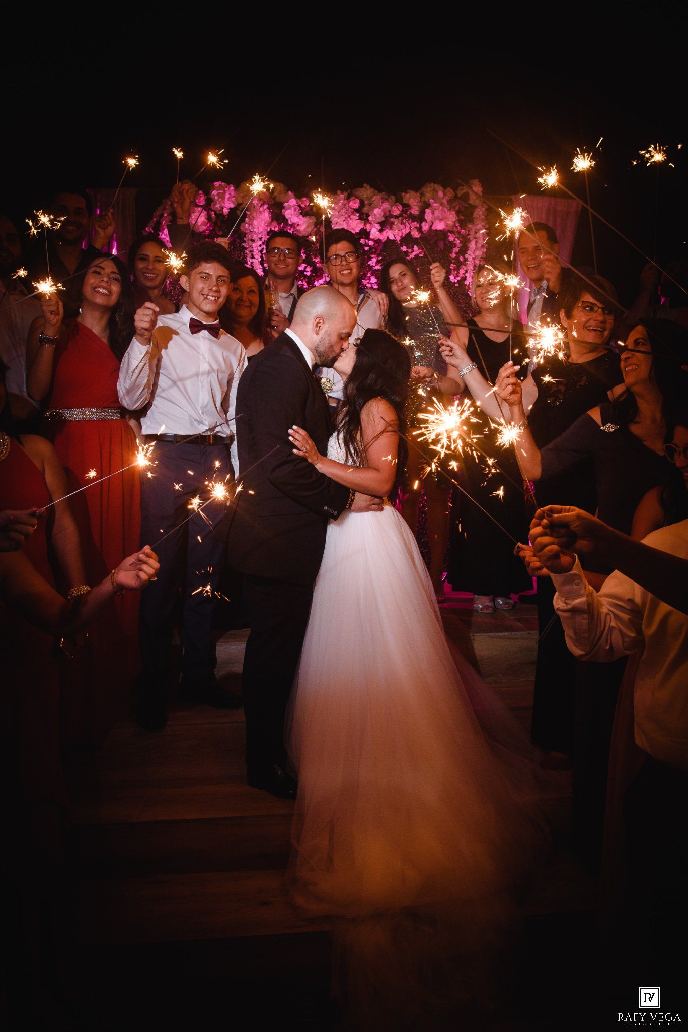 Bride and groom share a romantic kiss surrounded by wedding guests holding sparklers at a night reception.