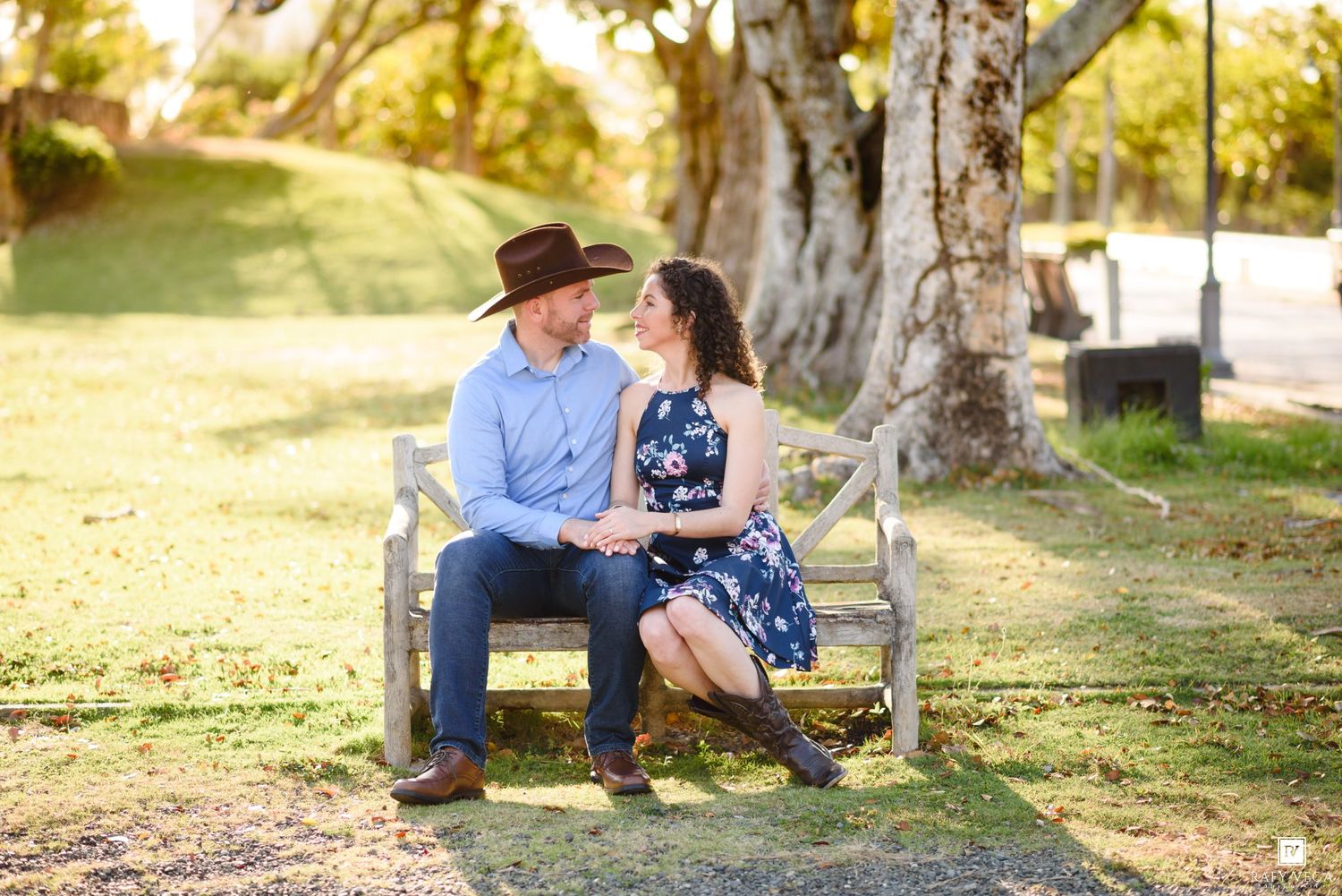 Couple sitting on a bench holding hands, smiling at each other in a sunny park with large trees.