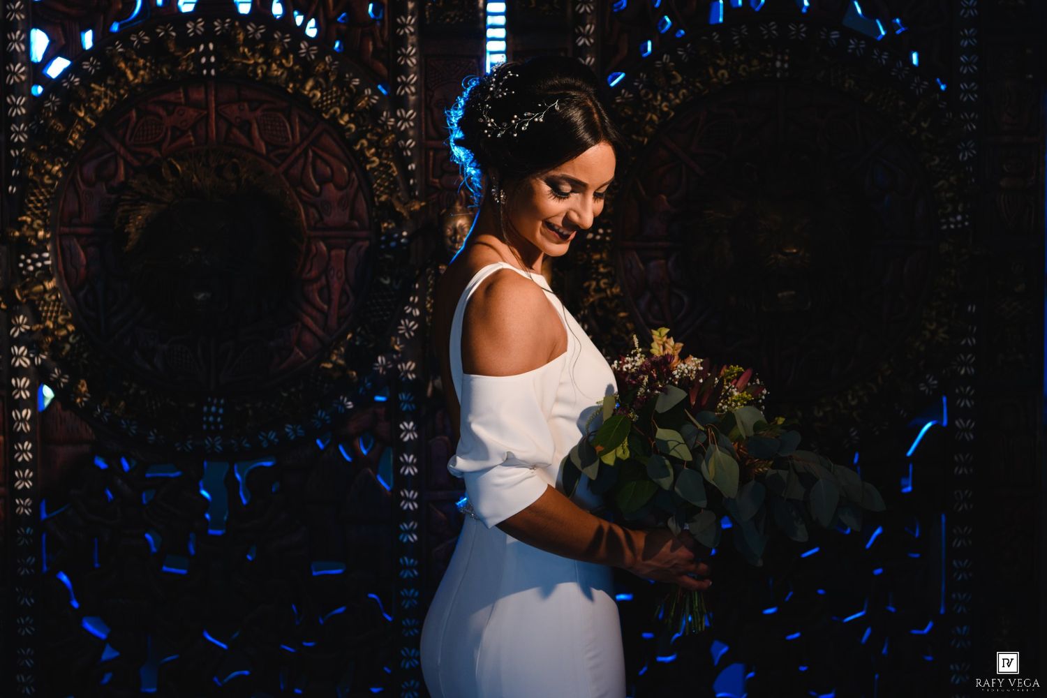 Bride in white off-shoulder gown smiling while holding a bouquet in a dark, ornate venue with blue lighting.