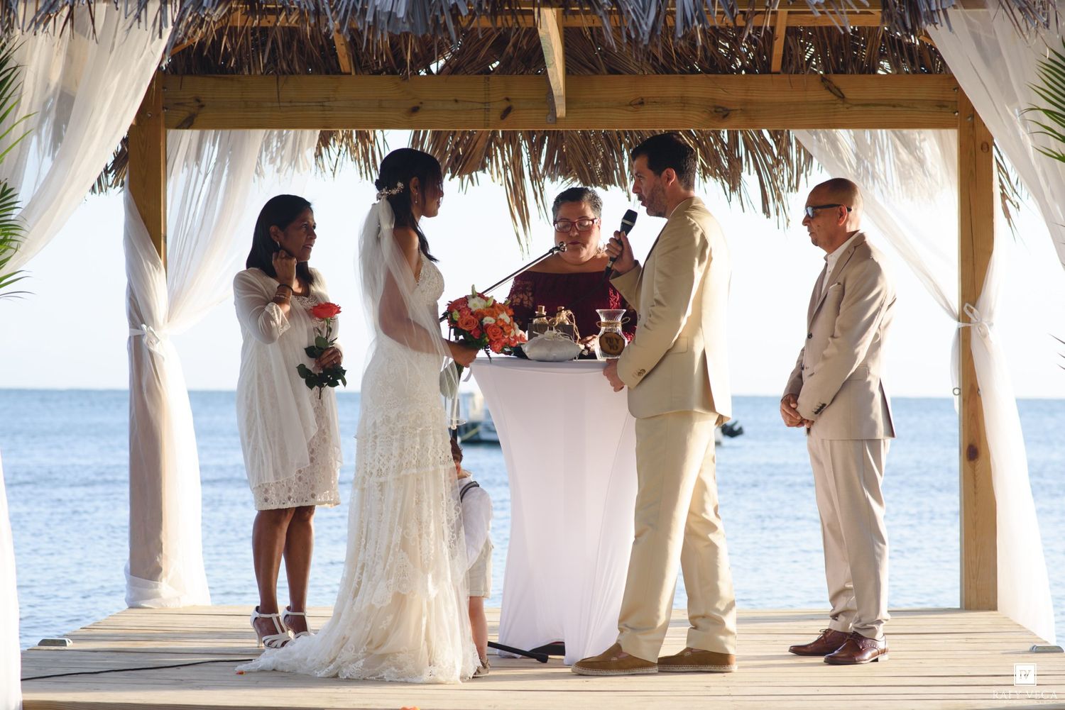 Couple exchanging vows at a beachside wedding ceremony under a thatched gazebo with ocean views.