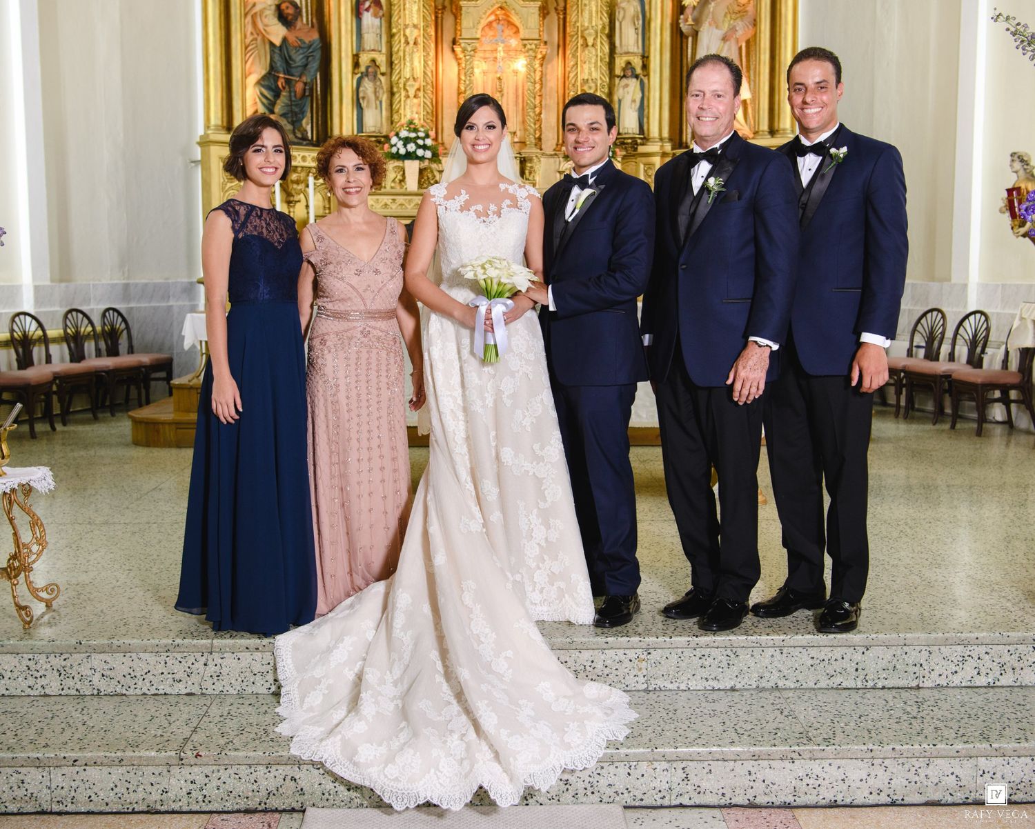 Wedding party poses in church, bride in lace gown with groom and family in navy formal attire.