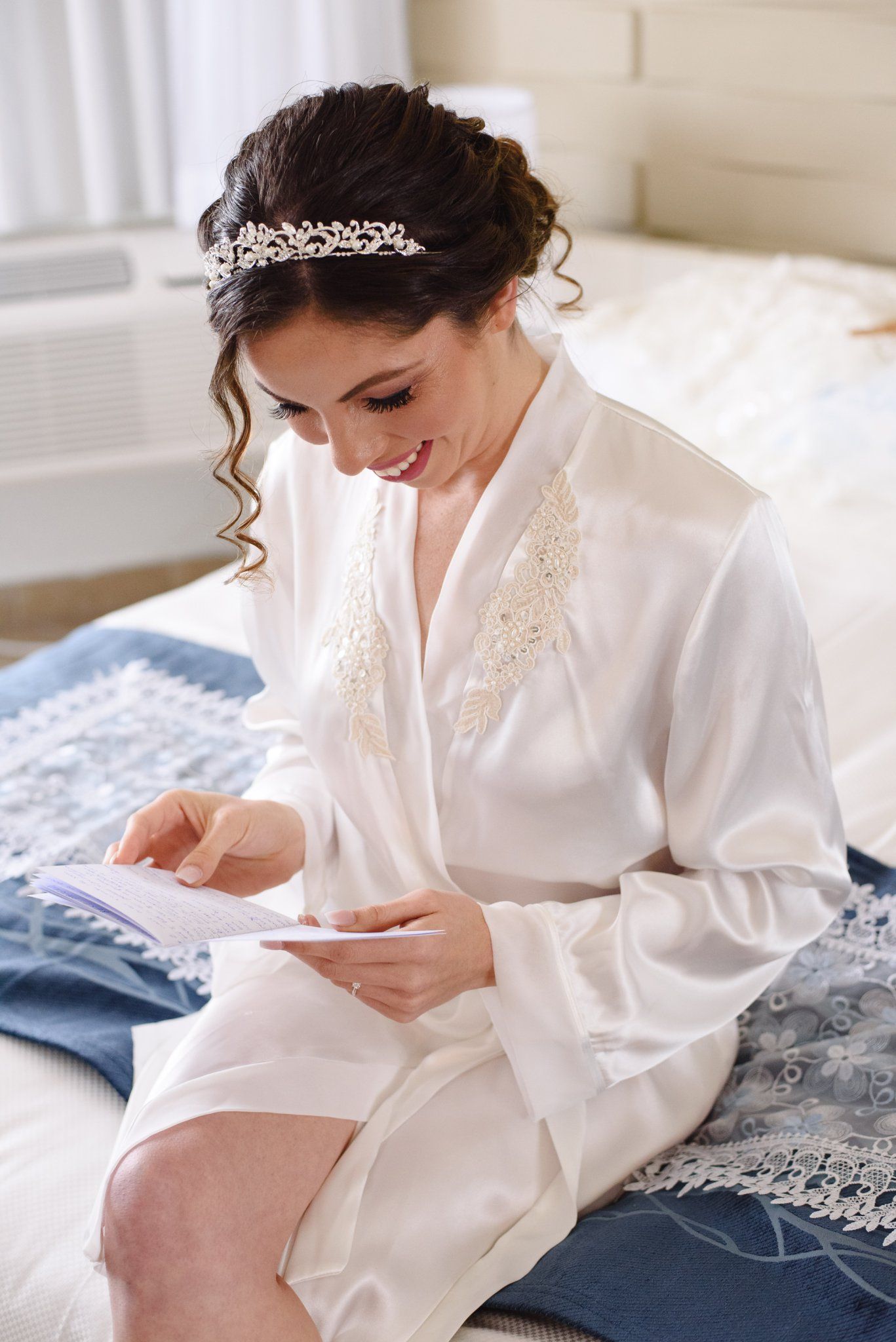 Smiling bride in white robe and crystal tiara reads a card while sitting on a bed before her wedding.