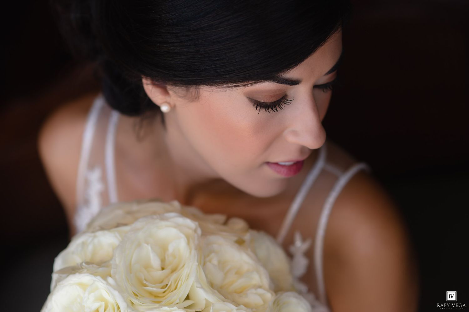 Elegant bride with dark updo holding white rose bouquet, wearing lace dress, soft bridal makeup, looking down gracefully.
