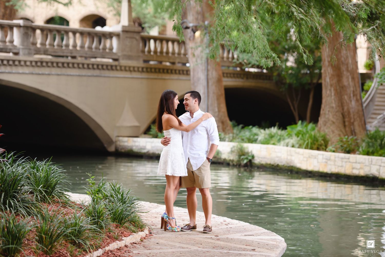 Couple embracing by the San Antonio River Walk with a stone bridge and lush greenery in the background.