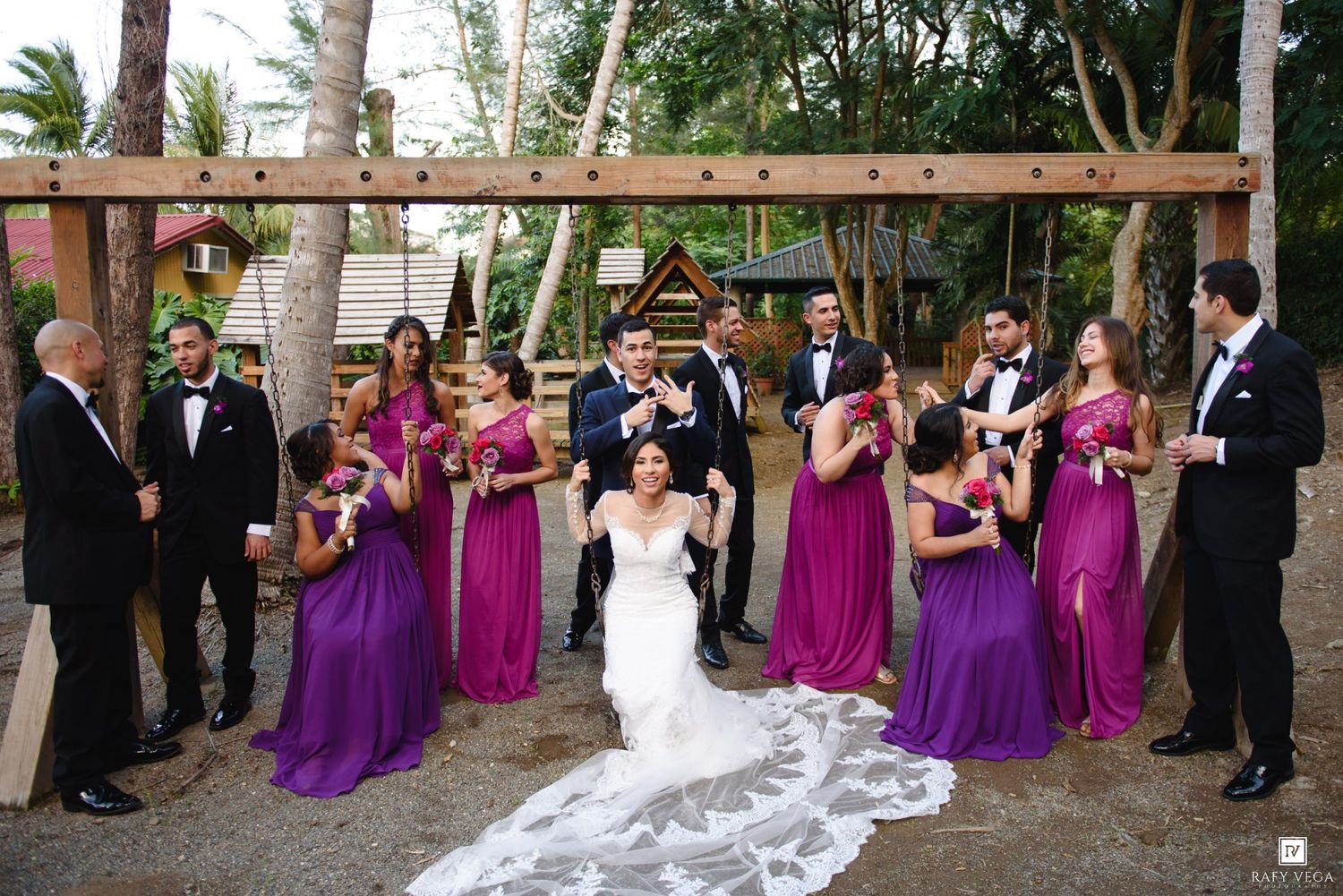 Wedding party poses outdoors with bridesmaids in purple gowns and groomsmen in tuxedos, bride and groom lifted by wedding party.
