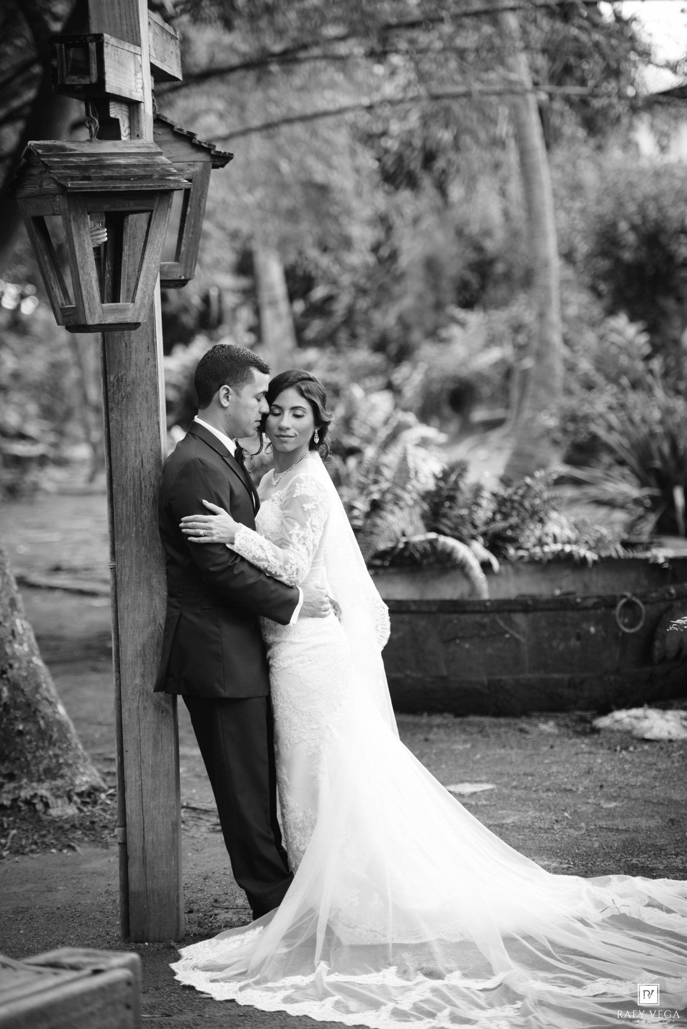 Black and white wedding photo of bride and groom embracing near a wooden lamp post in a lush garden setting.