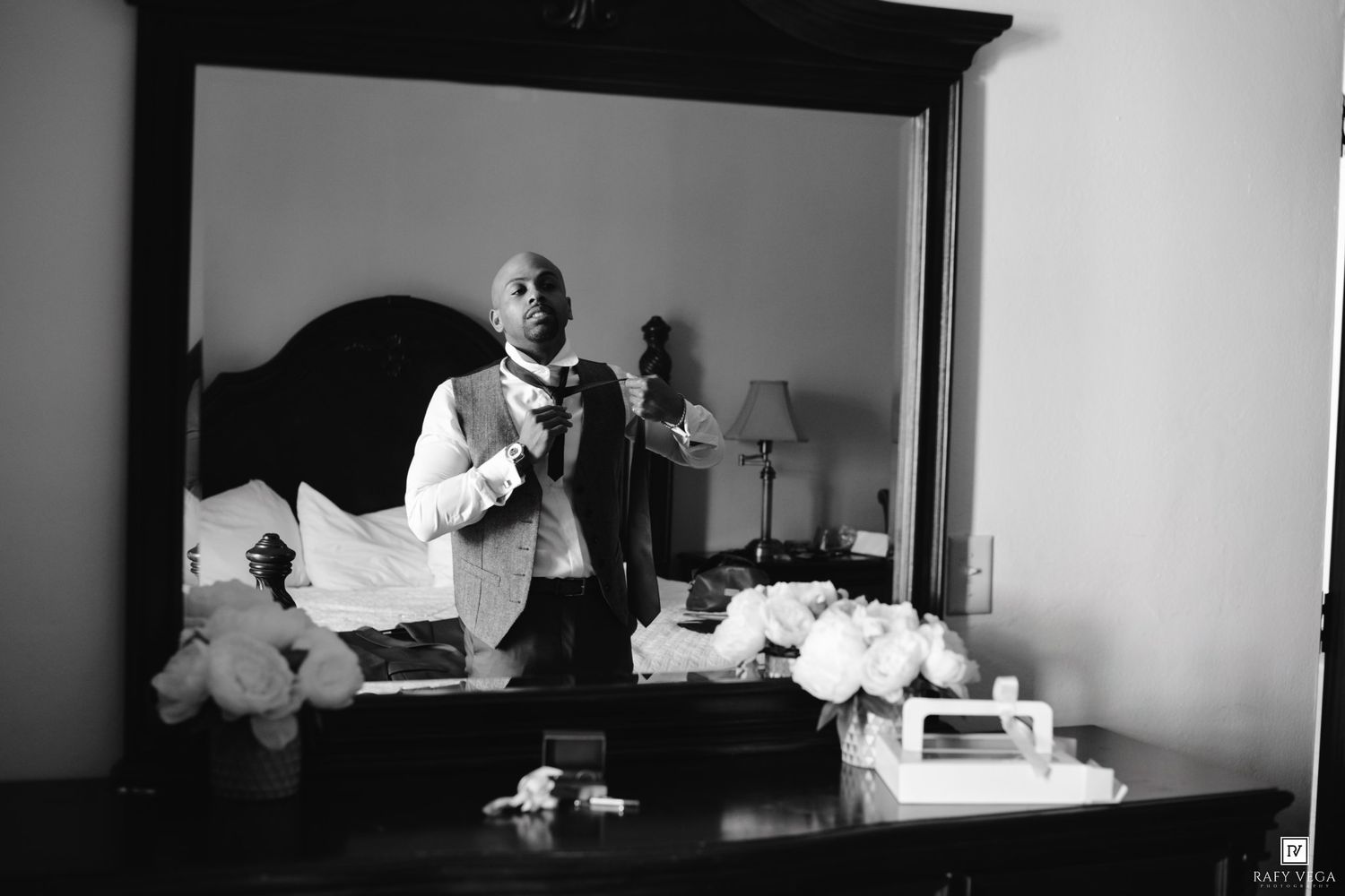 Black and white photo of a groom adjusting his tie in a hotel mirror while preparing for his wedding day.