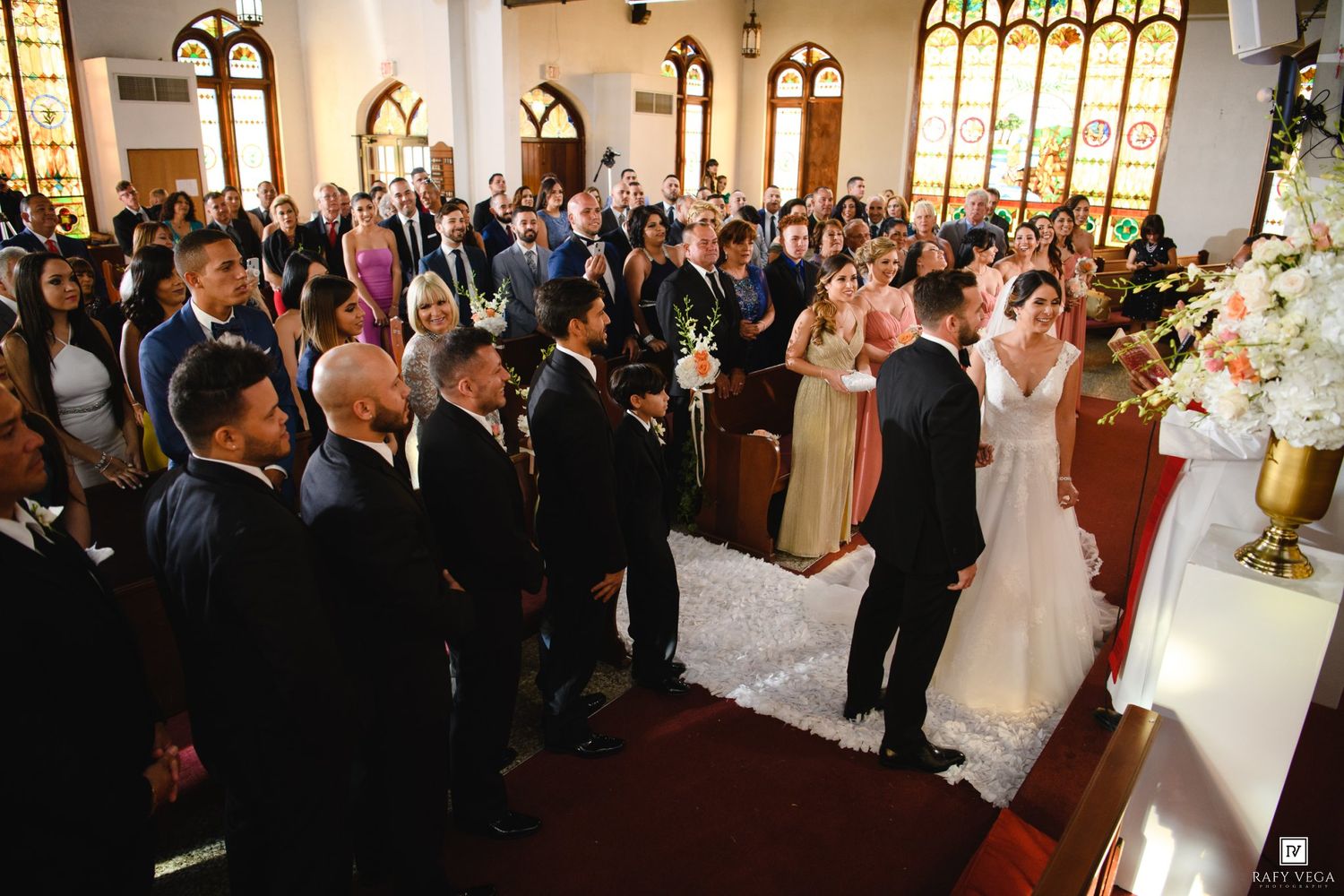 A bride and groom exchange vows at a church wedding ceremony surrounded by guests standing in decorated pews.