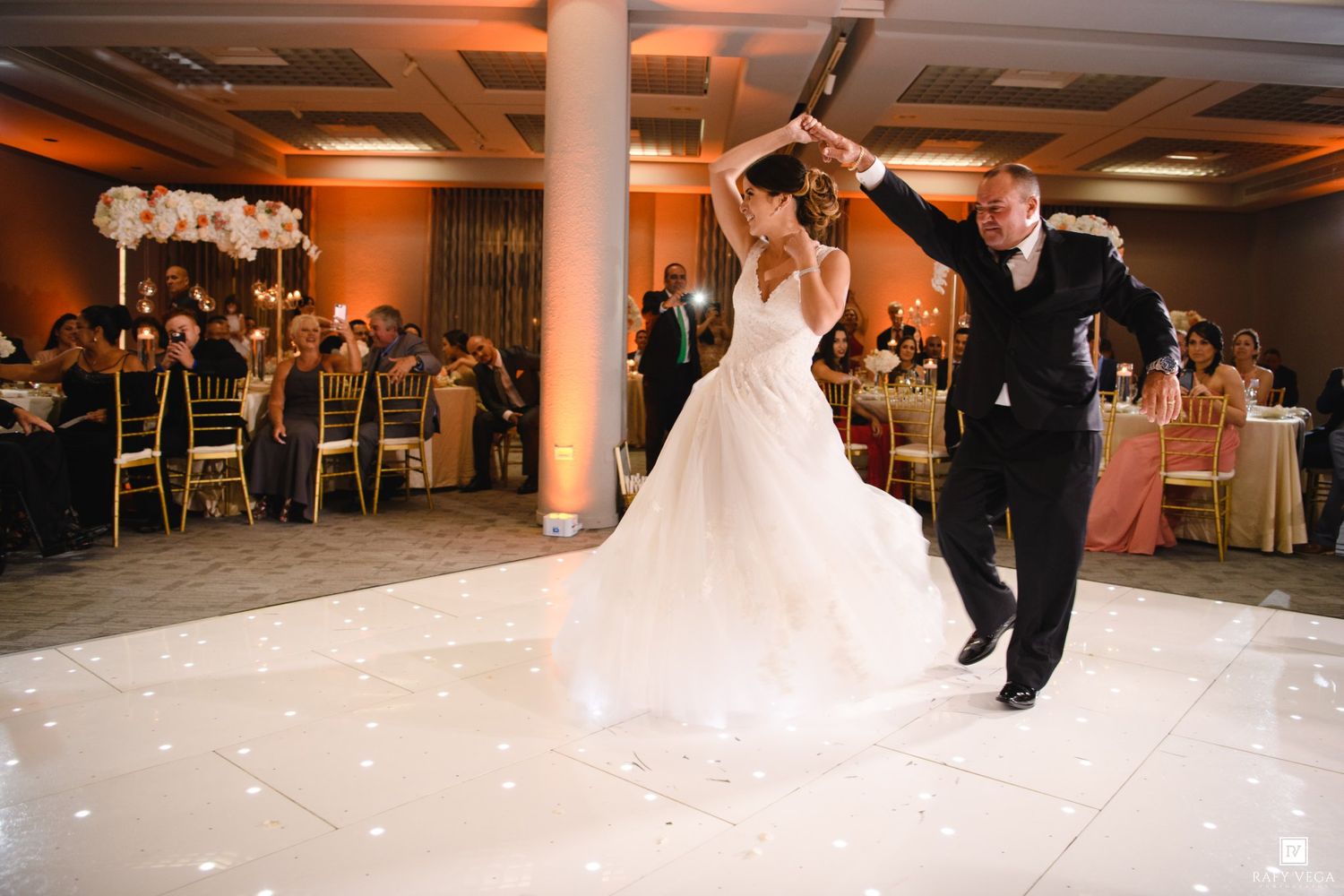 Bride and groom share first dance on white dance floor at elegant wedding reception with floral decor.