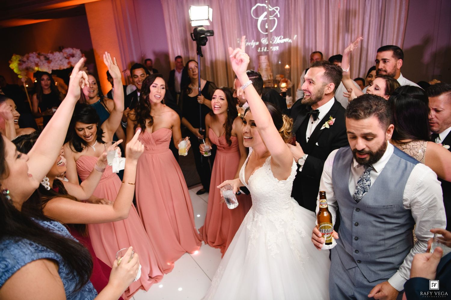 Bride and groom celebrate joyfully on the wedding dance floor surrounded by bridesmaids in pink dresses and cheering guests.
