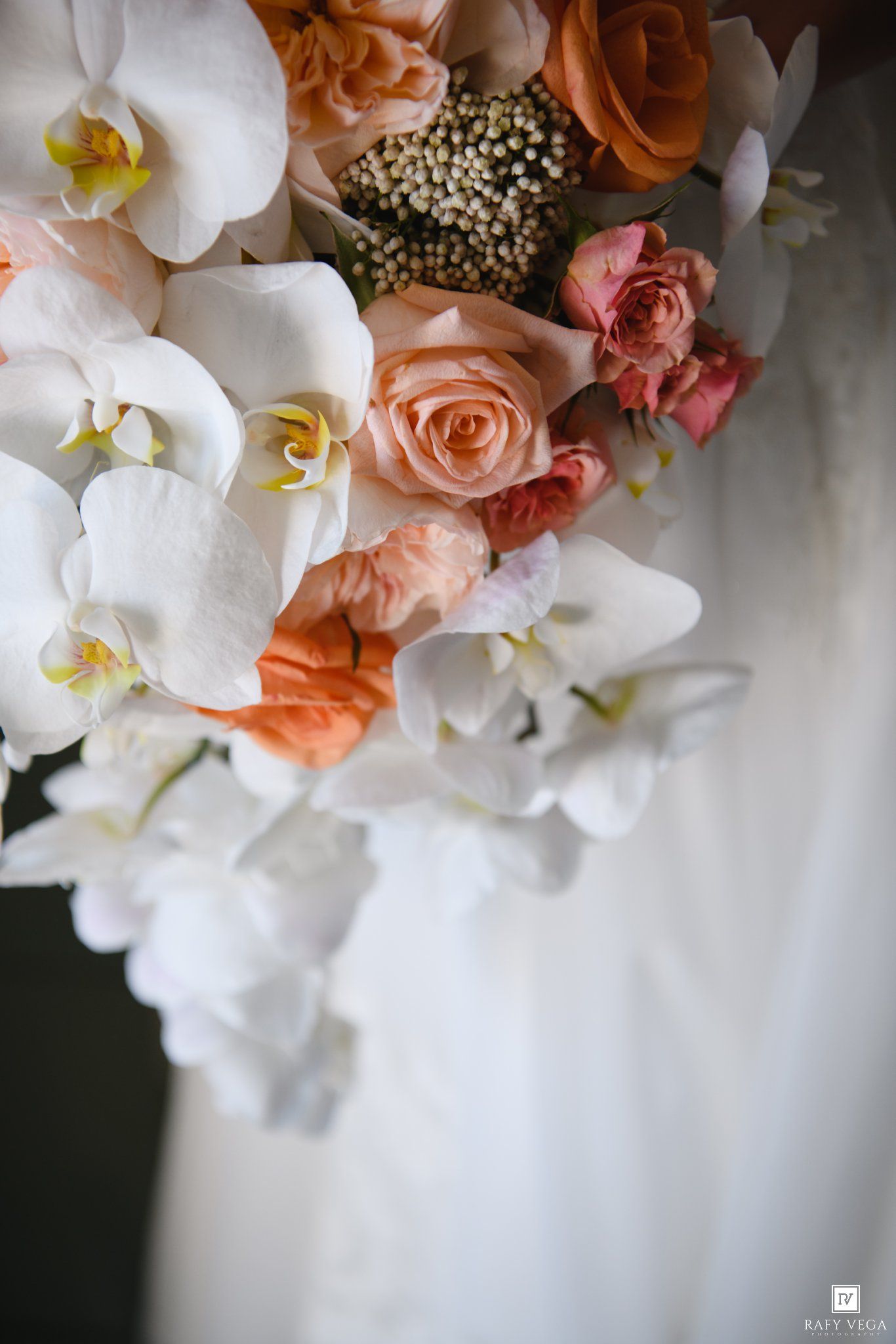 Bridal bouquet featuring white orchids, peach roses, coral flowers, and pearl accents against a soft white backdrop.
