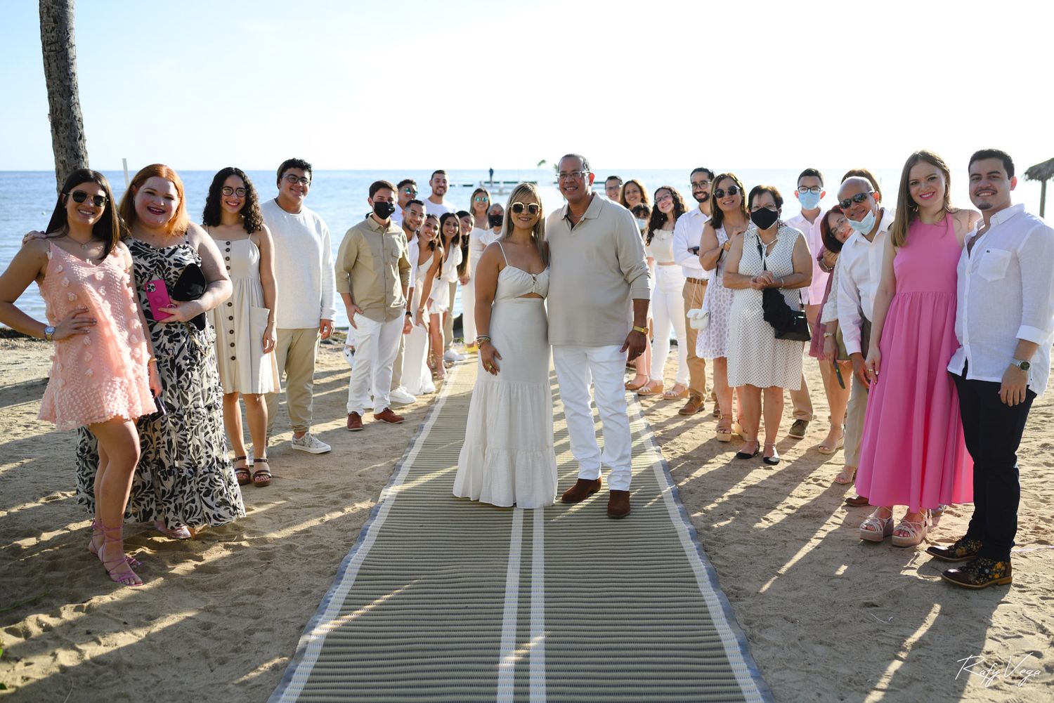 Bride and groom pose with wedding guests on a sunny beach, most dressed in white attire.