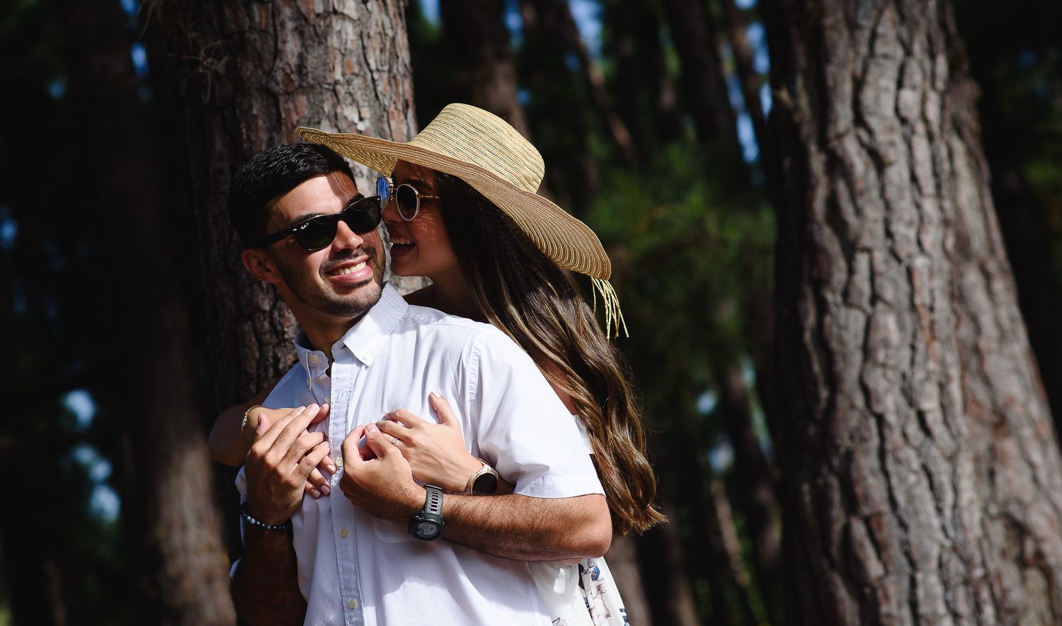 Couple embracing by a tree in a sunny forest, man in sunglasses and woman in a wide-brim straw hat.