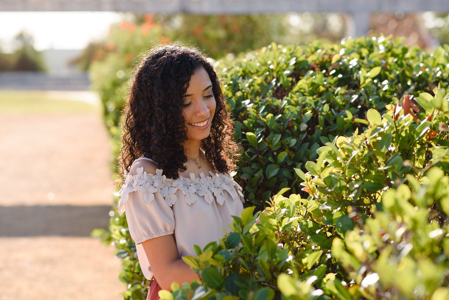 A smiling young woman with curly hair poses beside lush green hedges in a sunny outdoor garden setting.