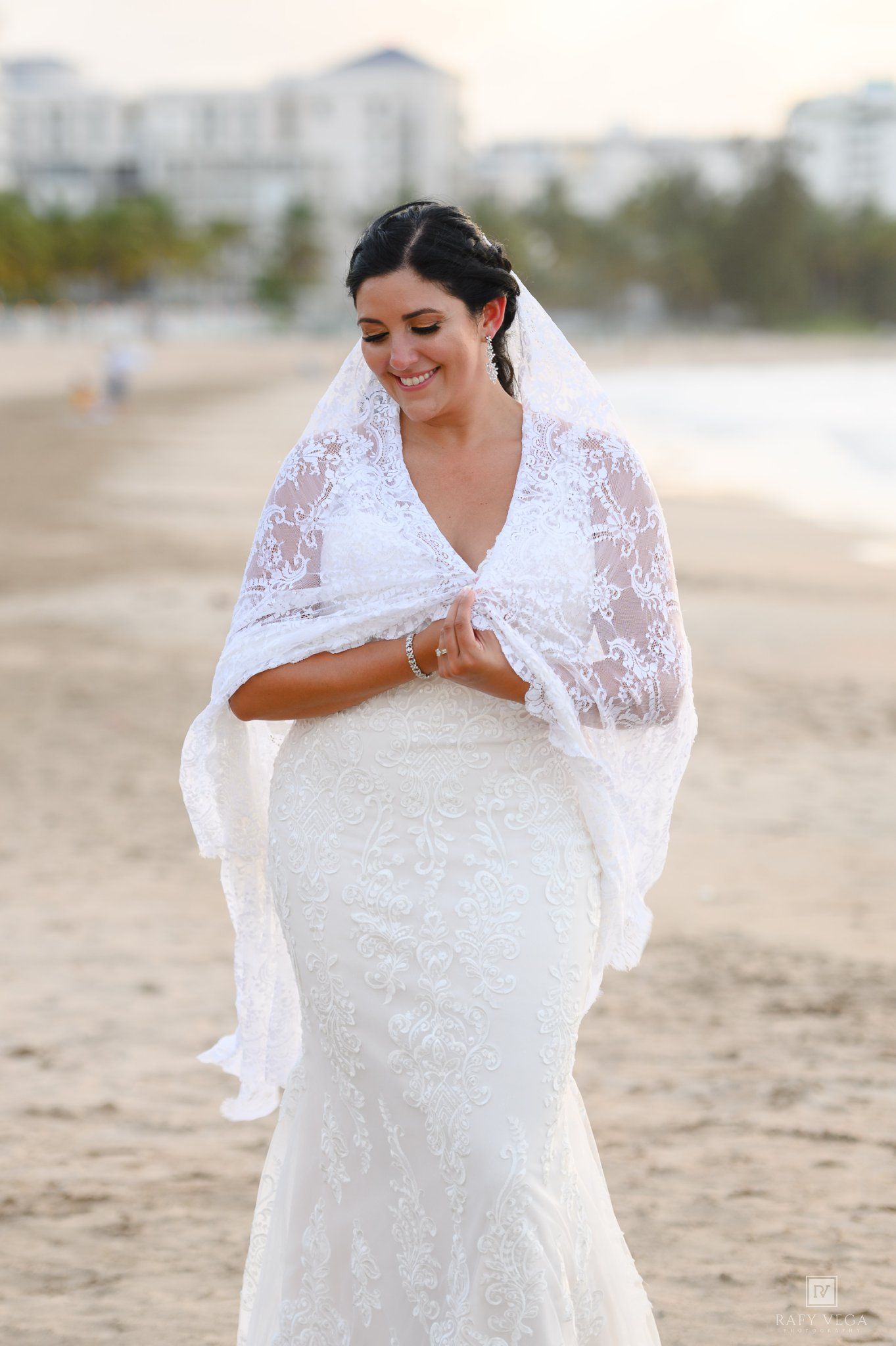 Bride wearing a lace mermaid wedding gown and matching lace shawl smiles softly on a sandy beach at sunset.