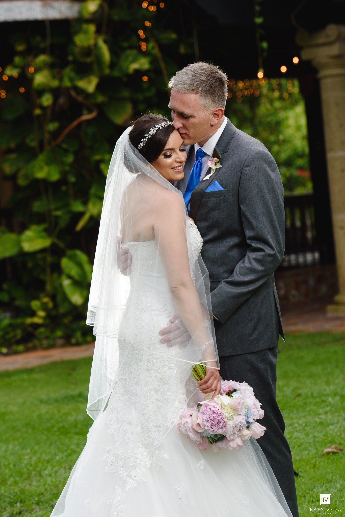 Bride in white lace gown with veil holds pink bouquet as groom in gray suit kisses her forehead outdoors.