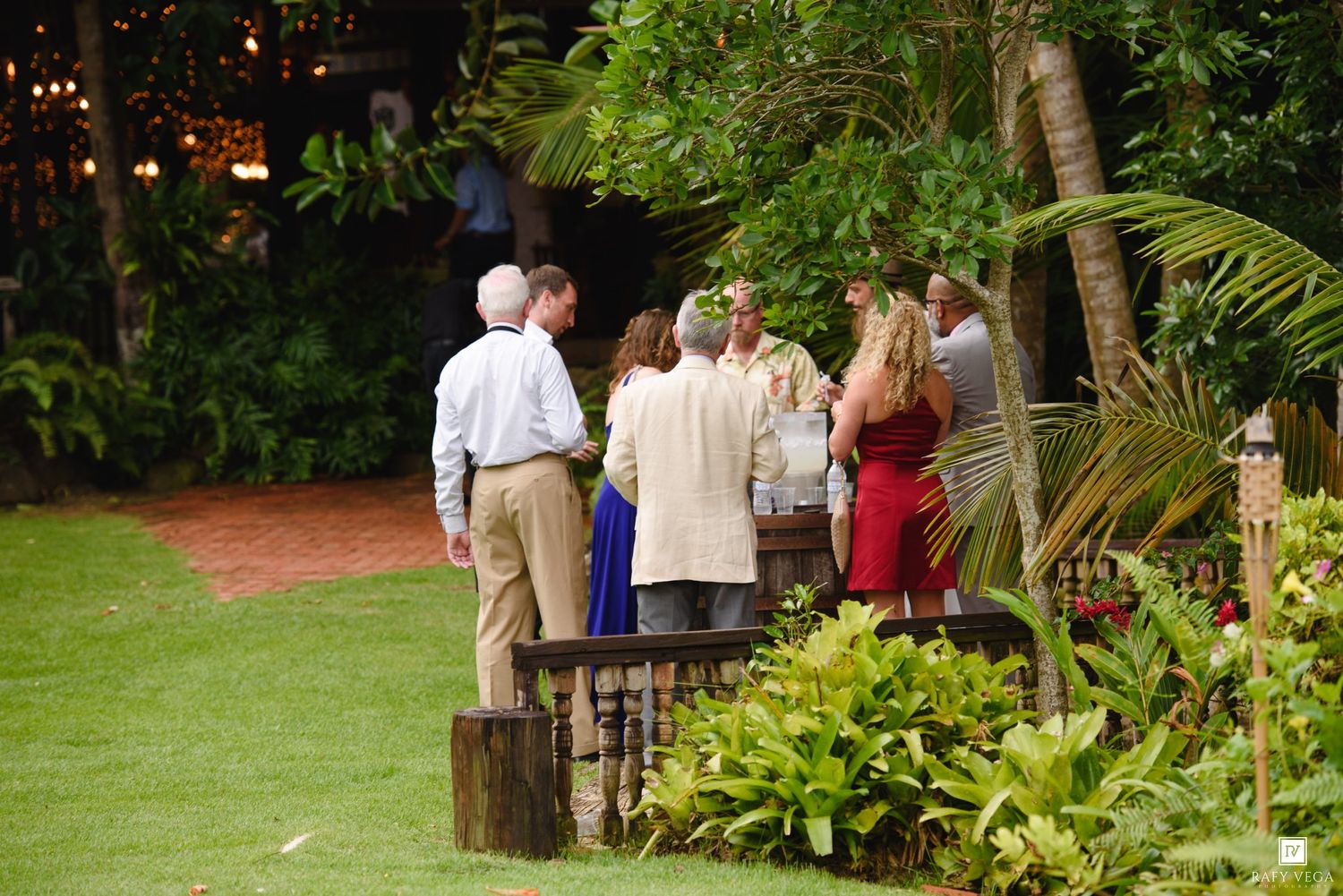 Group of elegantly dressed guests mingling outdoors at a tropical garden party surrounded by lush greenery.