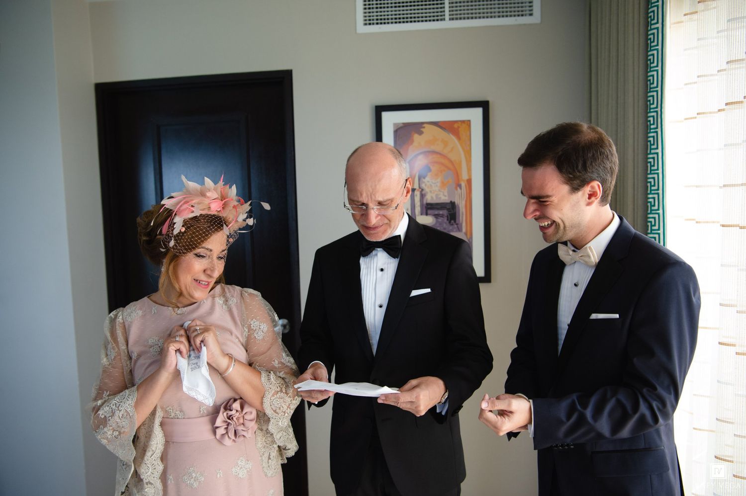 Three elegantly dressed people reviewing documents together in a bright room, woman in pink dress with floral headpiece.