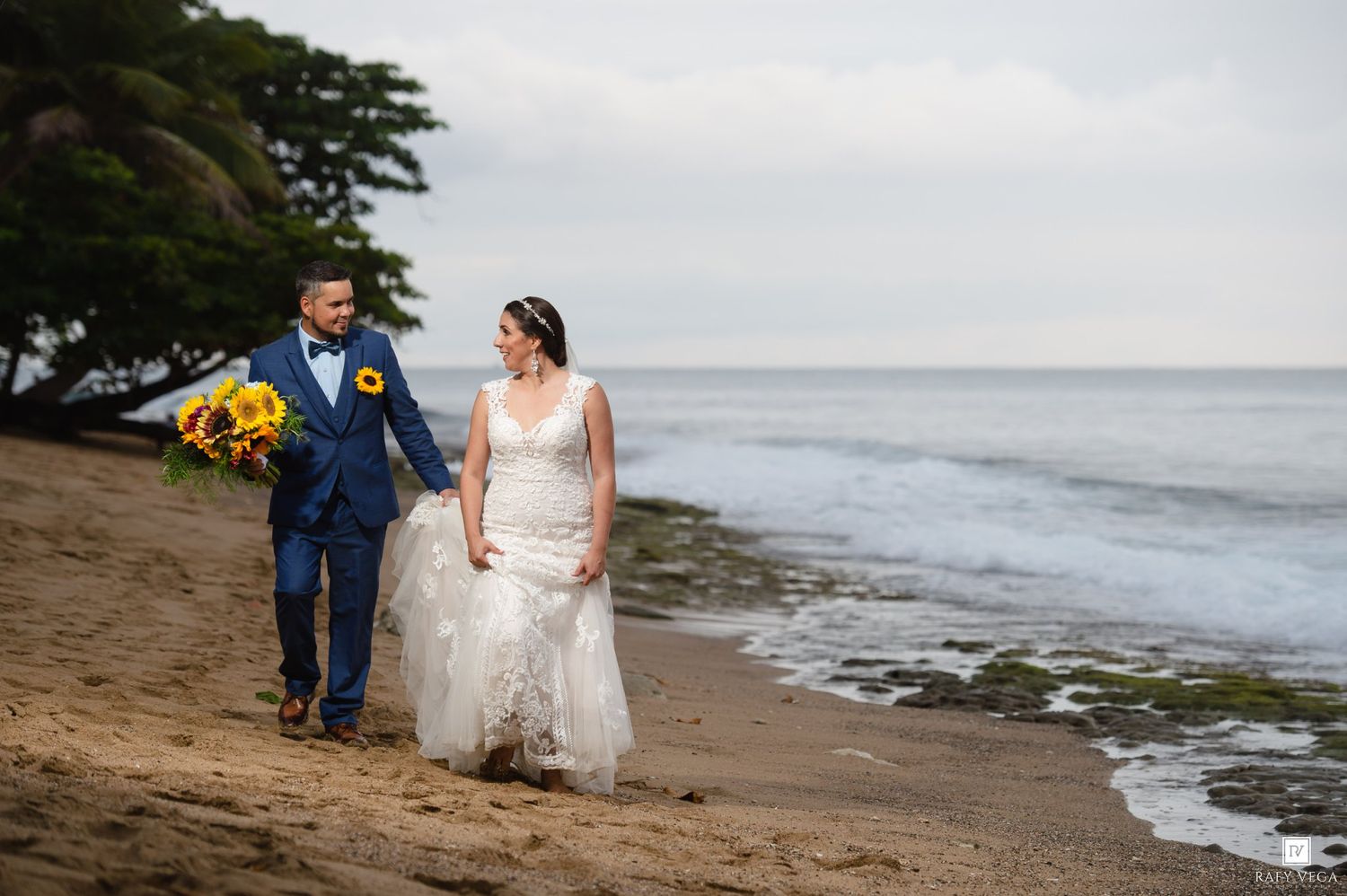 Bride and groom walk hand-in-hand on a sandy beach, she in lace gown, he in navy suit holding sunflowers.