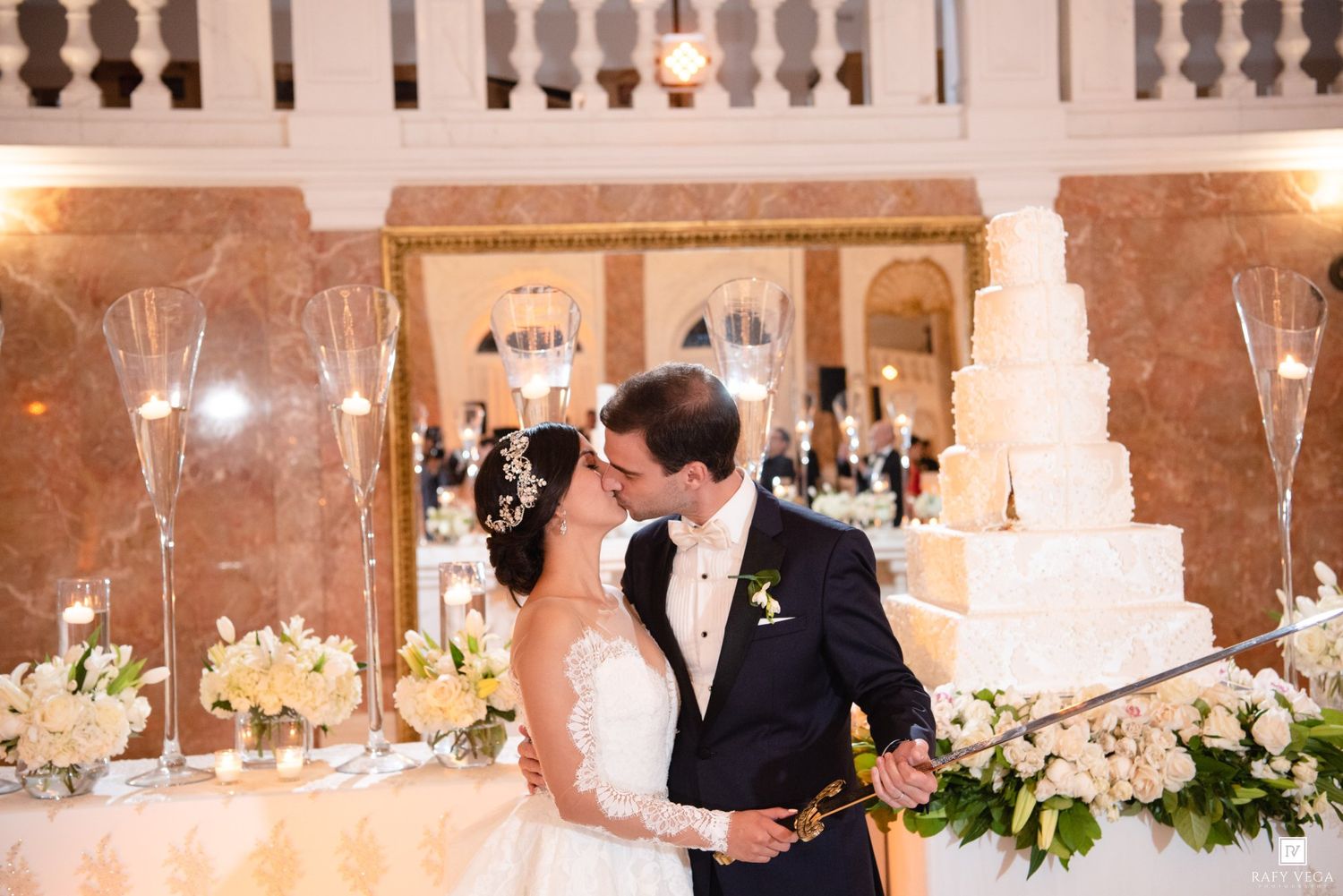 Bride and groom share a romantic kiss beside their elegant white wedding cake at a luxurious ballroom reception.