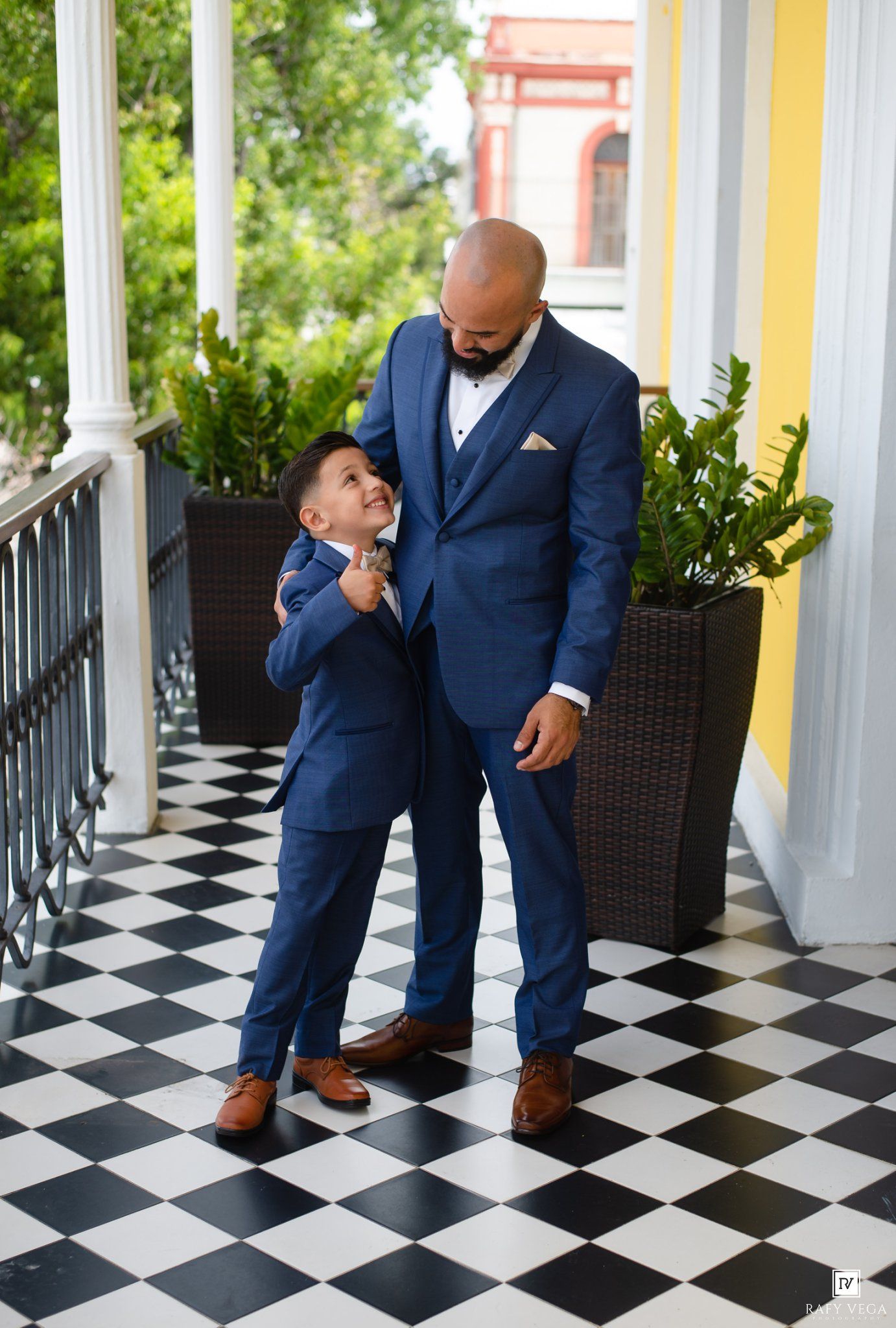 A man and young boy in matching blue suits stand on a checkered floor porch, sharing a sweet moment.
