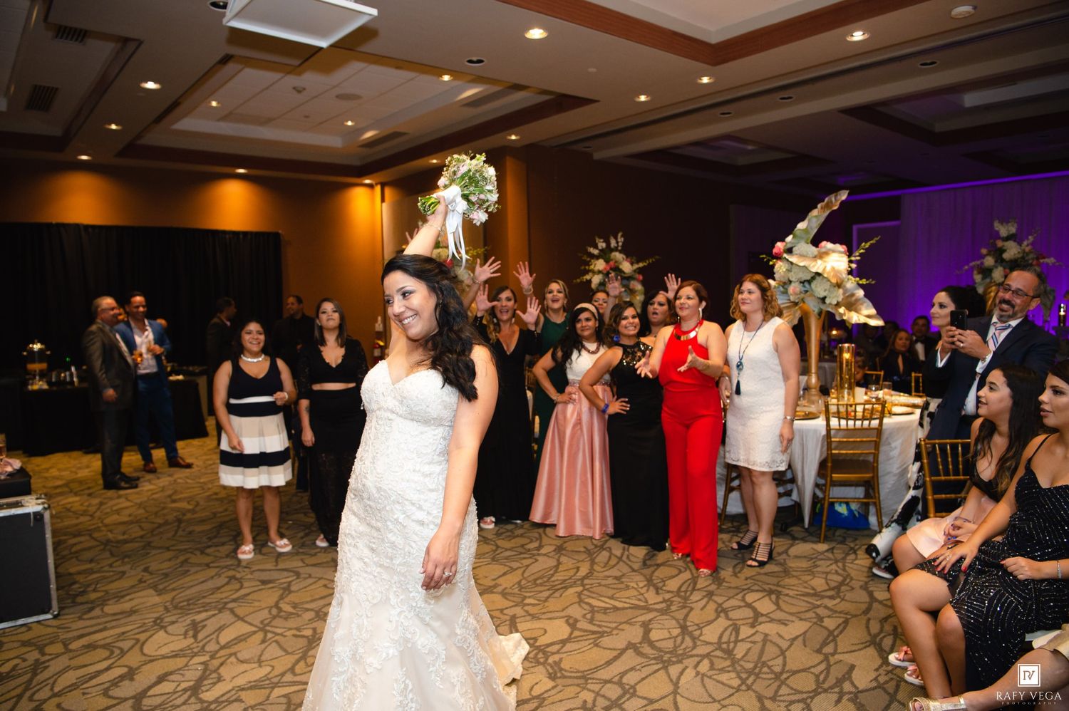 Bride in white wedding gown tossing bouquet to excited guests at elegant indoor wedding reception.