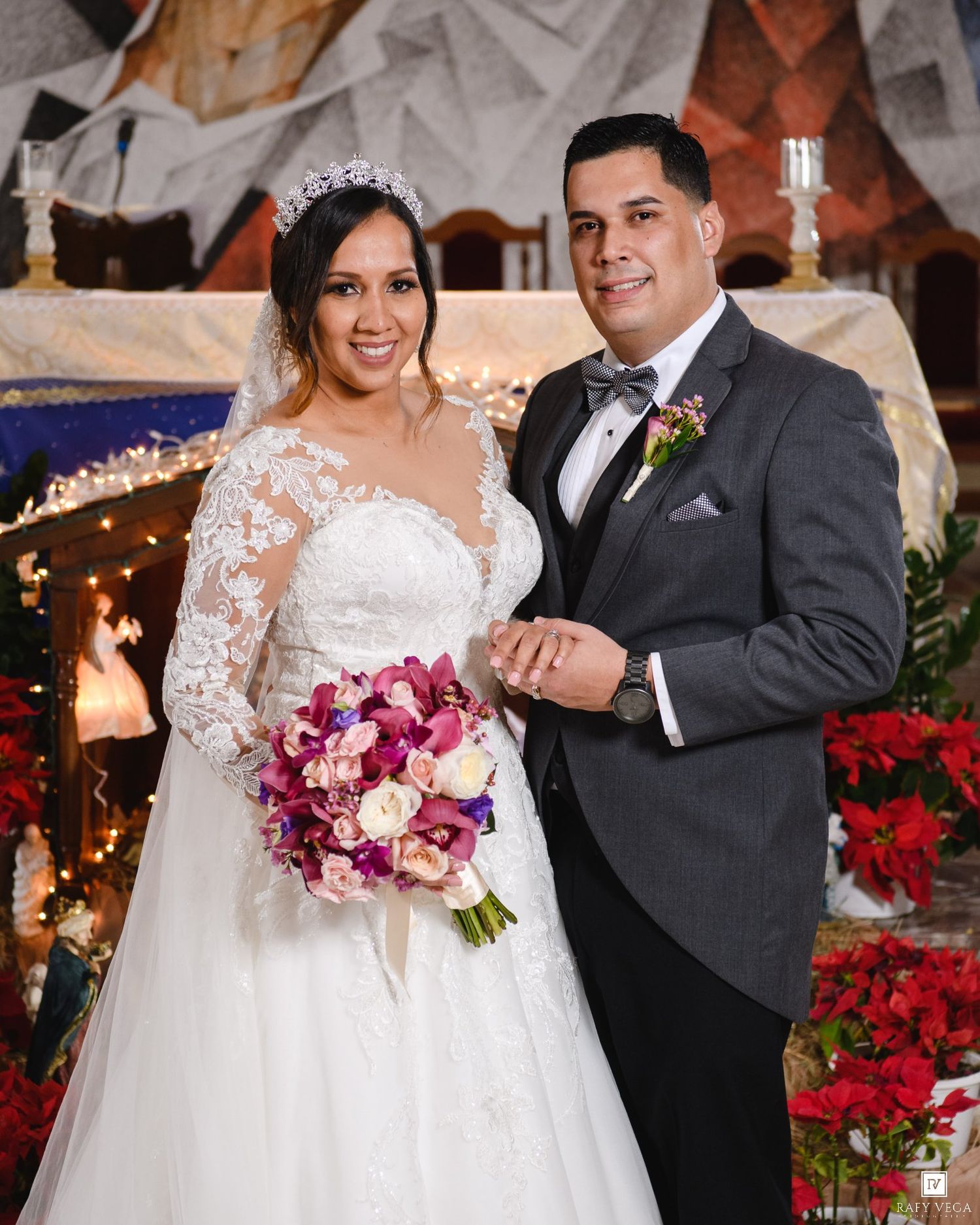 Bride and groom posing together at church altar, bride in lace gown with floral bouquet, groom in gray tuxedo.
