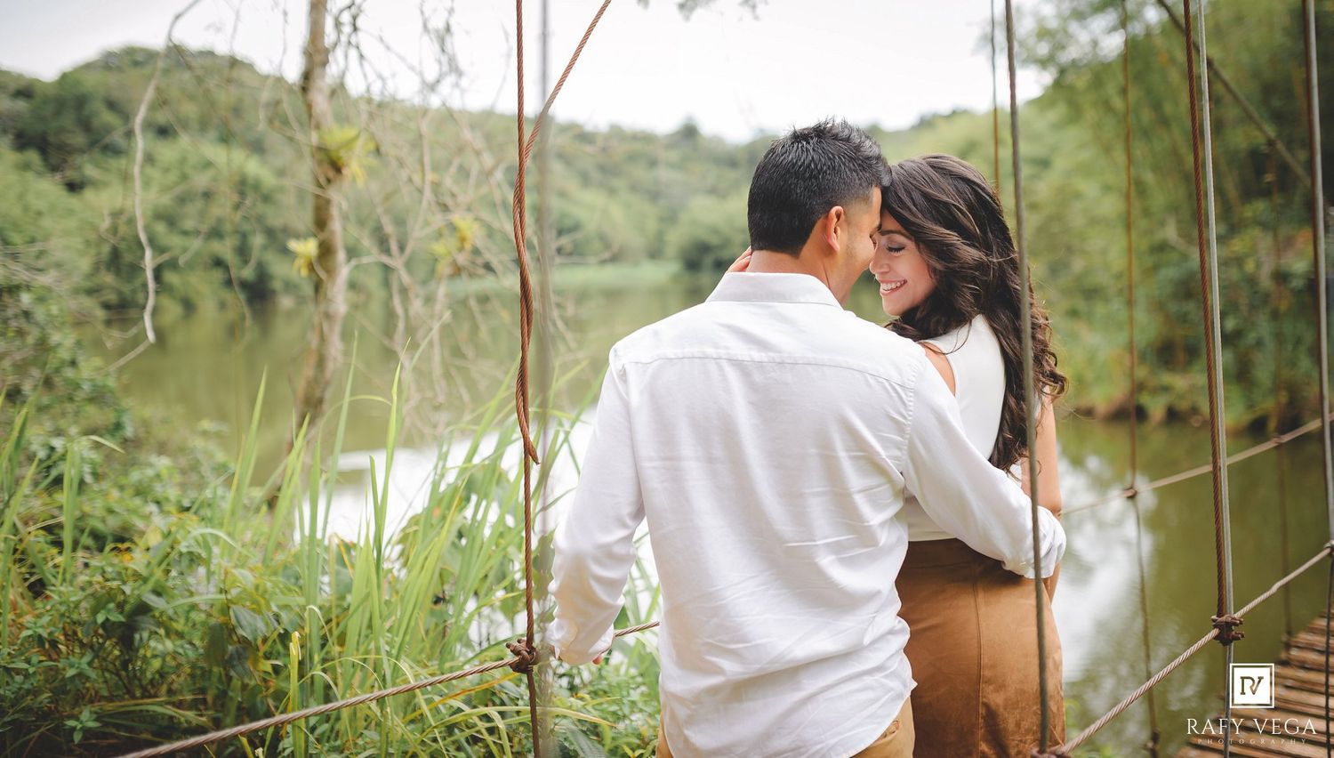 A couple embraces on a bridge surrounded by lush greenery and a serene river in a romantic outdoor setting.