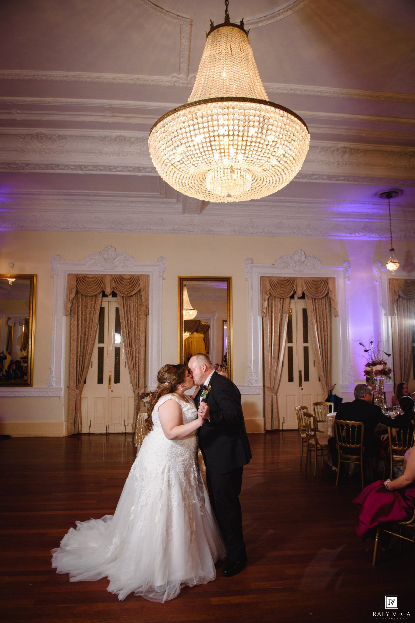 Bride and groom share first dance under crystal chandelier in elegant ballroom with gold drapes and purple uplighting.