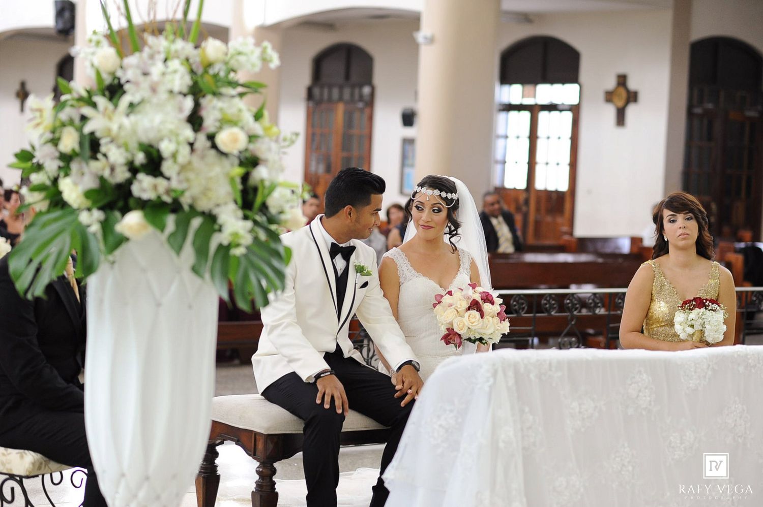 Bride and groom sitting together at church altar during wedding ceremony with white floral arrangements.