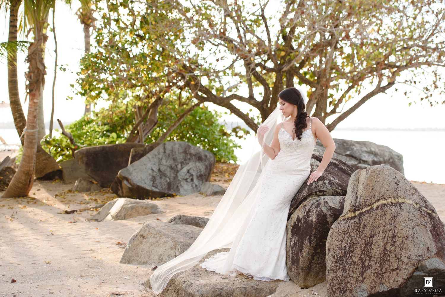 Bride in white wedding gown sitting on rocks at a tropical beach with palm trees and sand in the background.