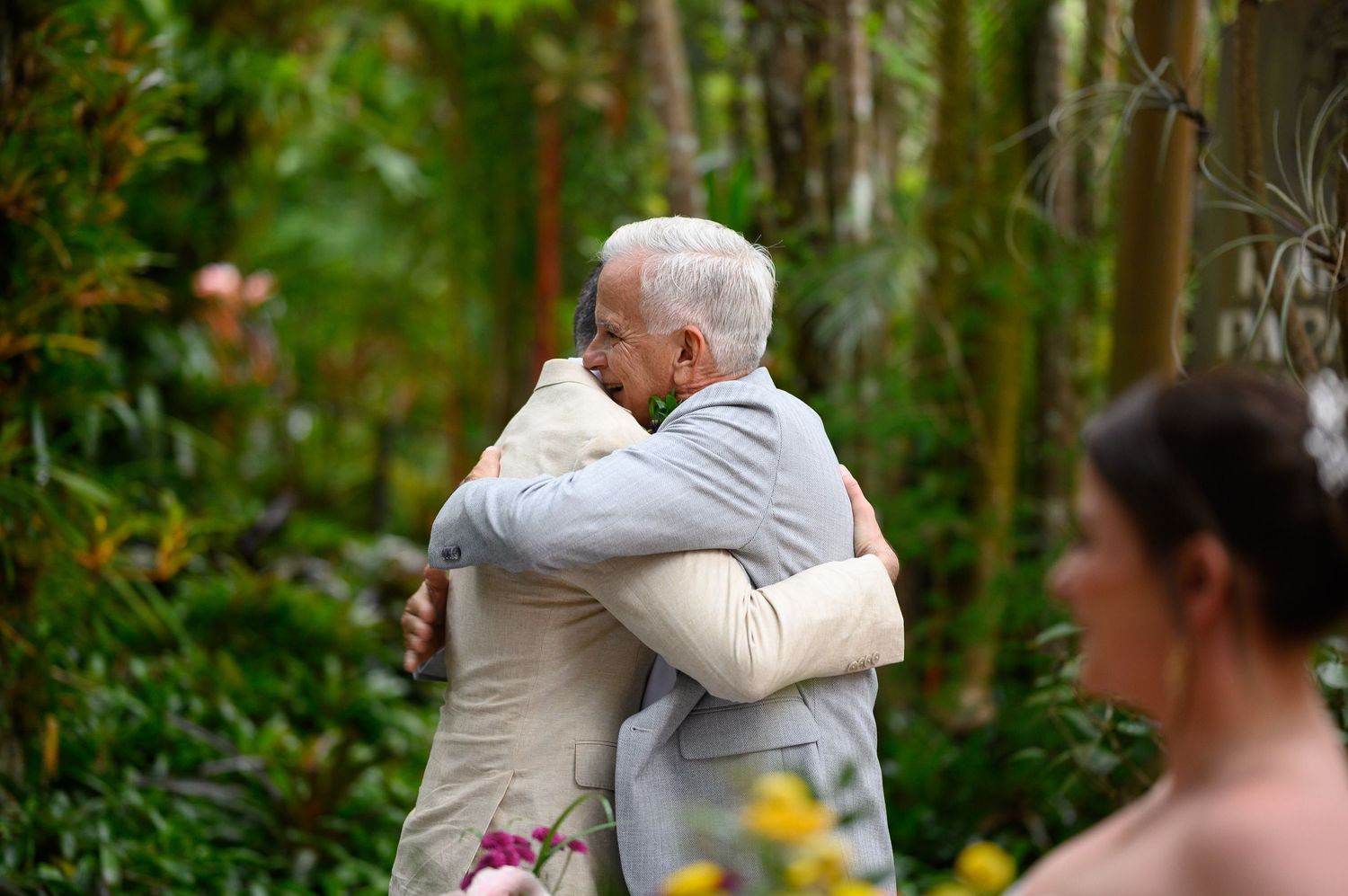 Two people share a warm embrace at an outdoor wedding ceremony surrounded by lush tropical greenery.