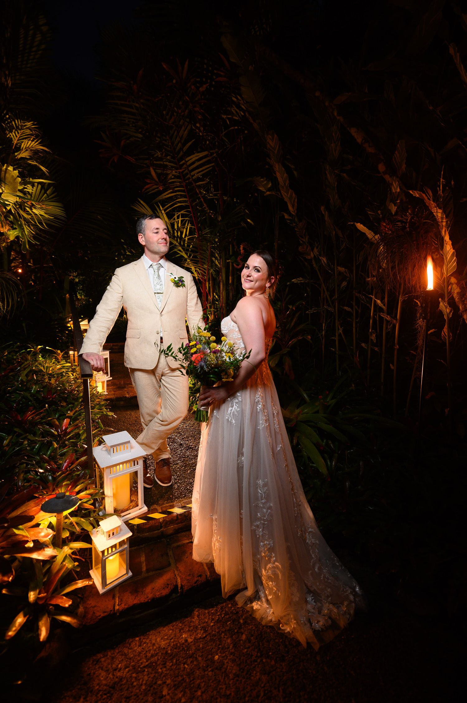 Bride and groom pose at night in tropical garden with lanterns, she holds colorful bouquet in lace gown.