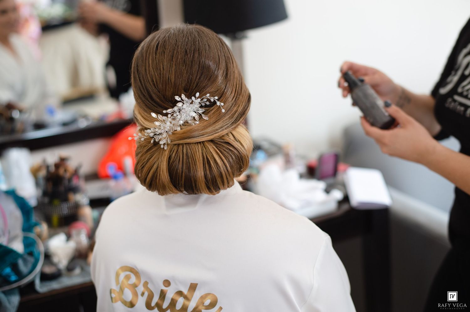 Bride in white robe with elegant updo hairstyle adorned with floral hair accessory being styled by hairdresser.