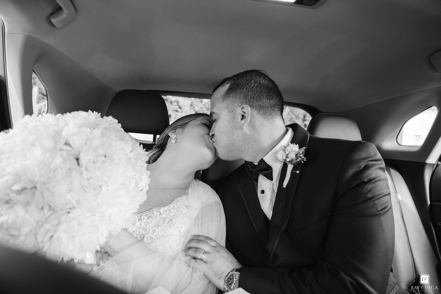 Black and white photo of a bride and groom sharing a kiss in the back seat of a car on their wedding day.