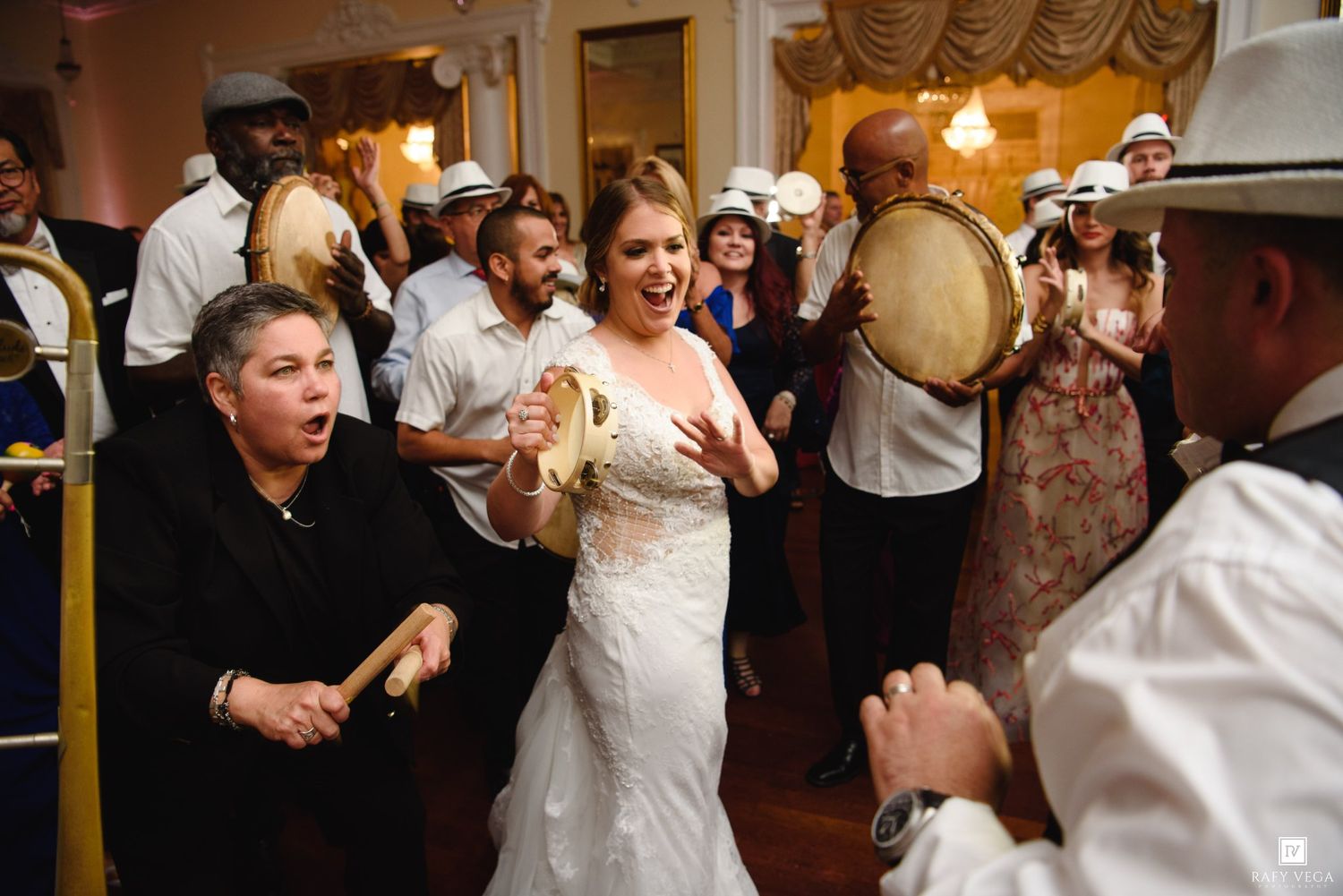 Bride in white gown dancing joyfully with band musicians playing tambourines at lively wedding reception.